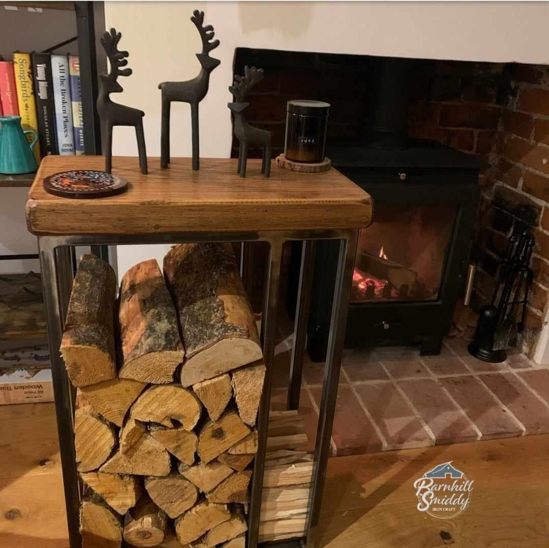 The image shows a wooden topped log holder table with a side panel for kindling positioned next to a fireplace. Here's a breakdown of th elements in the image: Log holder table - the main focus is a table with a wood top and a metal frame which is buffed down and lacquered with clear lacuer tp keep the raw steel look and protect it. The frame forms a rectangular section underneath the table top which is filled with neatly stacked logs. A smaller section to the side, also frame in metal, holds kindling.